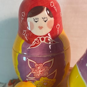 May include: A ceramic nesting doll featuring a red headscarf with white floral outlines, a purple apron with a large pink and yellow flower design, and a bright yellow body. The doll has a painted face with closed eyes and pink cheeks.
