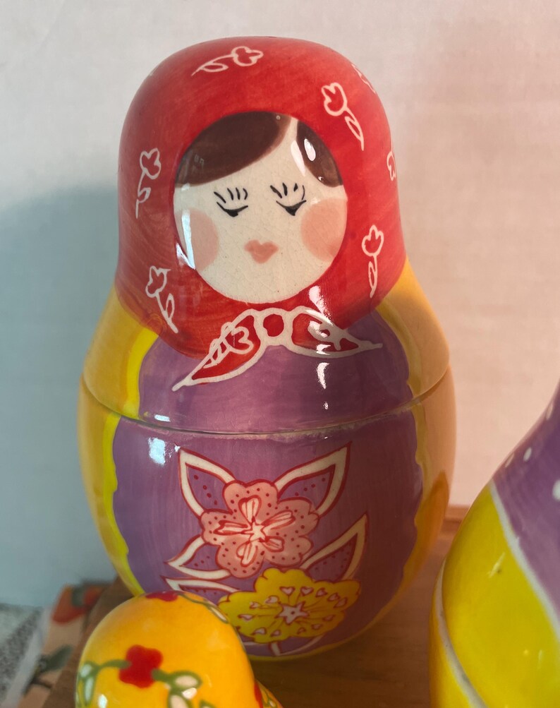 May include: A ceramic nesting doll featuring a red headscarf with white floral outlines, a purple apron with a large pink and yellow flower design, and a bright yellow body. The doll has a painted face with closed eyes and pink cheeks.