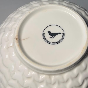May include: White ceramic bowl with a textured, quilted pattern on the exterior. The bottom of the bowl is white with a black bird logo inside a circle, and the text "MADE IN CHINA FABRIQU&Eacute; EN CHINE".