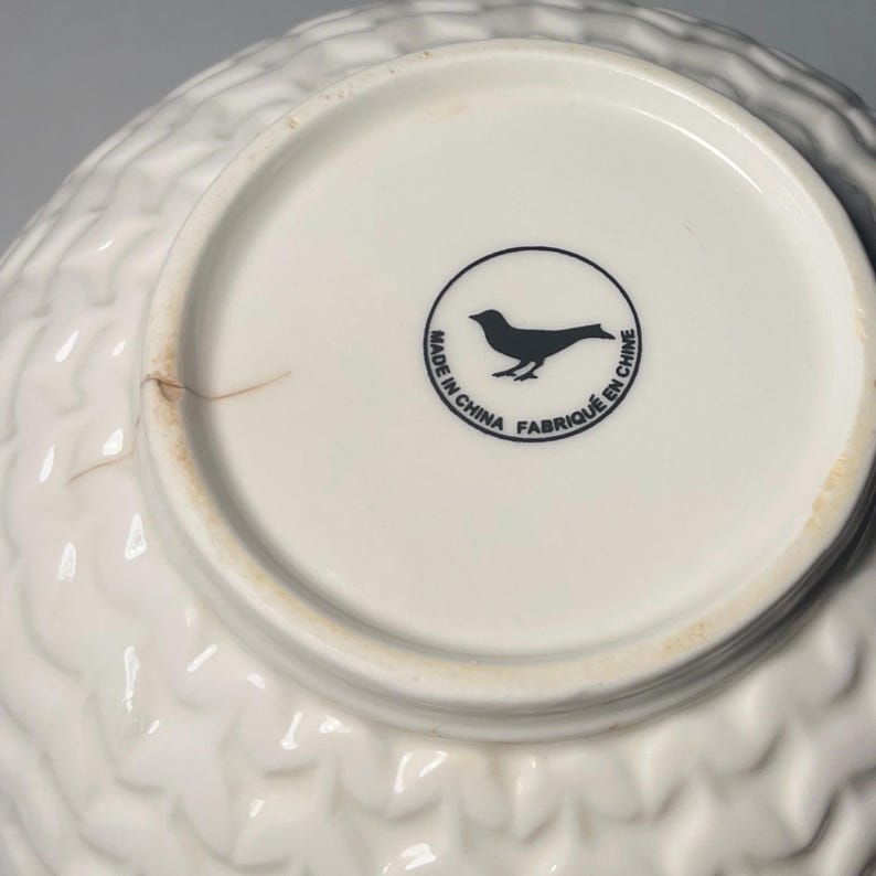 May include: White ceramic bowl with a textured, quilted pattern on the exterior. The bottom of the bowl is white with a black bird logo inside a circle, and the text "MADE IN CHINA FABRIQU&Eacute; EN CHINE".