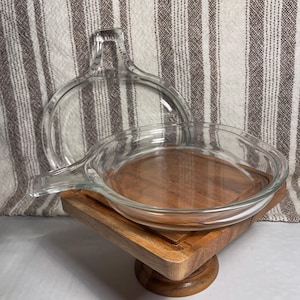 May include: A clear glass baking dish with a handle and a matching lid. The dish is sitting on a wooden stand. The background is a striped fabric.