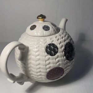 May include: A white ceramic teapot with a textured surface and a lid. The teapot features decorative circular patterns in black and burgundy. The lid is topped with a gold-colored knob. The teapot has a curved handle and a spout.