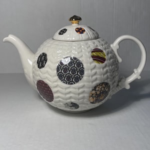 May include: White ceramic teapot with a round body and lid, adorned with patterned circles in various colors. The teapot features a curved spout, a handle, and a gold-colored knob on the lid. The body has a textured, wavy pattern.