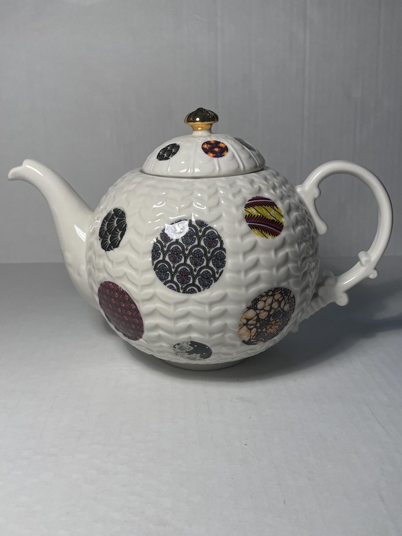 May include: White ceramic teapot with a round body and lid, adorned with patterned circles in various colors. The teapot features a curved spout, a handle, and a gold-colored knob on the lid. The body has a textured, wavy pattern.
