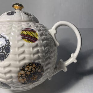 May include: White ceramic teapot with a textured surface and colorful patterned circles. The lid is topped with a gold-colored knob. The handle and spout are white.