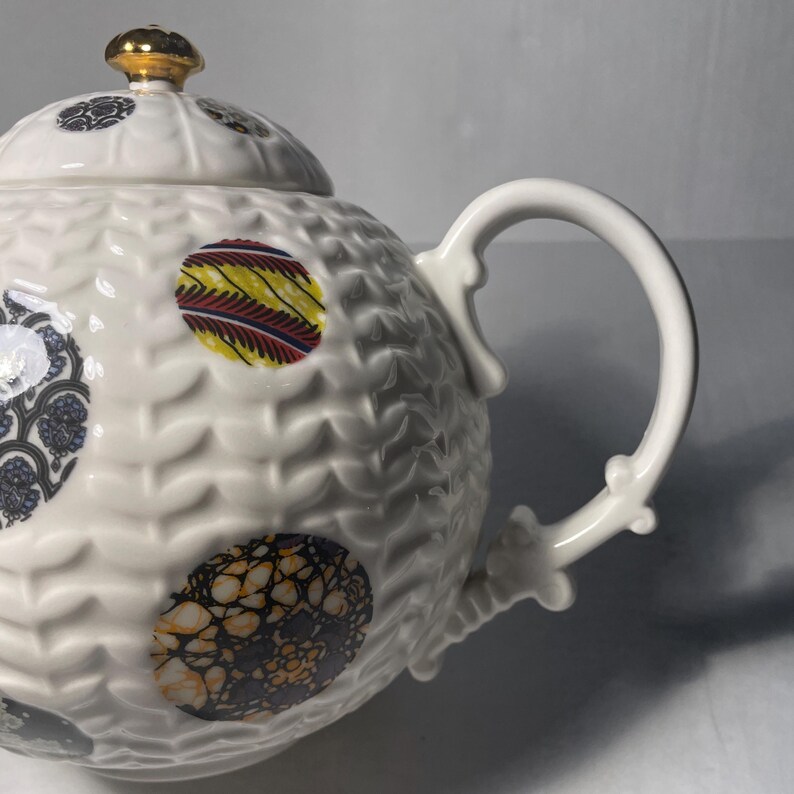 May include: White ceramic teapot with a textured surface and colorful patterned circles. The lid is topped with a gold-colored knob. The handle and spout are white.