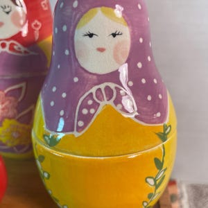 May include: A nesting doll featuring a purple headscarf with white polka dots, a yellow body, and a painted face with rosy cheeks. The doll has a smooth, glossy finish and is decorated with green vine patterns along the lower yellow section.