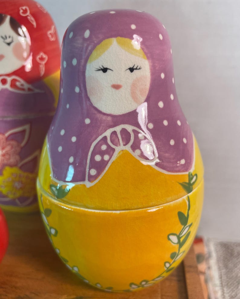 May include: A nesting doll featuring a purple headscarf with white polka dots, a yellow body, and a painted face with rosy cheeks. The doll has a smooth, glossy finish and is decorated with green vine patterns along the lower yellow section.