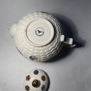 May include: White ceramic teapot with a textured, patterned body and a matching lid. The lid features a gold knob and decorative polka dots in various colors. The bottom of the teapot has a circular emblem with a bird silhouette.
