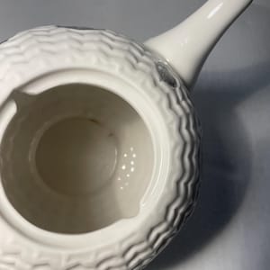 May include: A white ceramic teapot with a textured, wave-like pattern around the body. The spout and handle are smooth and white. The interior is a light beige color. There are small, dark gray accents on the body.