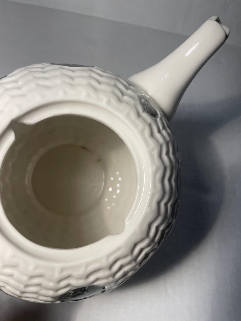 May include: A white ceramic teapot with a textured, wave-like pattern around the body. The spout and handle are smooth and white. The interior is a light beige color. There are small, dark gray accents on the body.