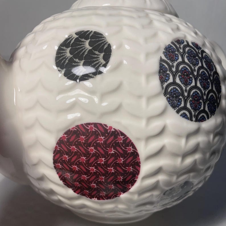 May include: White ceramic teapot with a textured surface and three patterned circular accents. The patterns include black and white, blue and red, and red and white designs. The teapot has a spout and a lid.