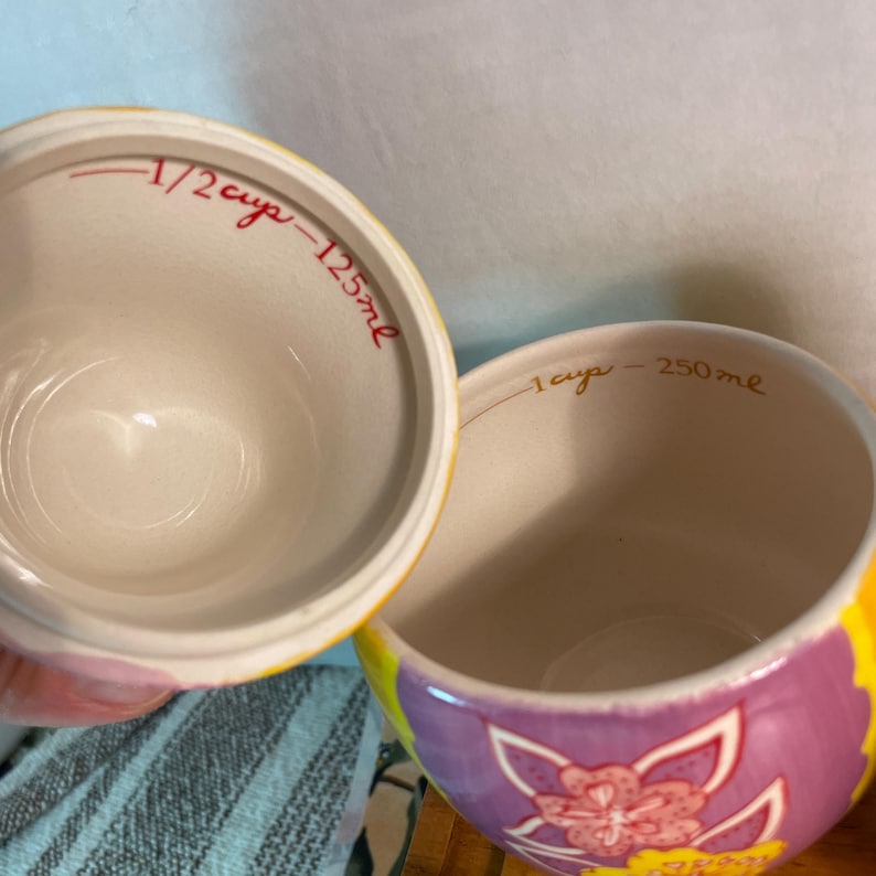 May include: Two ceramic measuring cups with colorful floral patterns on the exterior. The smaller cup interior is marked 1/2 cup - 125ml in red text, while the larger cup interior is marked 1 cup - 250ml in gold text. Both have smooth, off-white interiors.