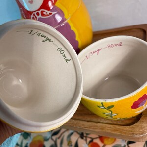 May include: Two ceramic measuring cups with a yellow exterior and floral pattern. The interior of the left cup is inscribed with 1/4 cup and 60ml, while the right cup features an inscription of 1/3 cup and 80ml in a handwritten script style.