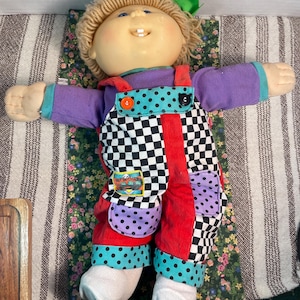 May include: A doll with blonde yarn hair and a green hair bow, wearing a purple long-sleeve shirt under checkered and red corduroy overalls. The overalls feature two purple polka-dot pockets, one orange button, one black button, and a small patch.
