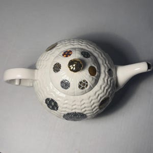 May include: White ceramic teapot with a textured surface and a lid adorned with patterned circles in various colors. The teapot features a gold knob and a curved spout, perfect for serving tea.