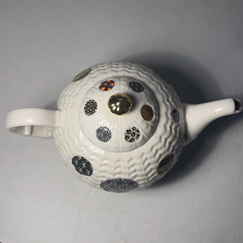 May include: White ceramic teapot with a textured surface and a lid adorned with patterned circles in various colors. The teapot features a gold knob and a curved spout, perfect for serving tea.