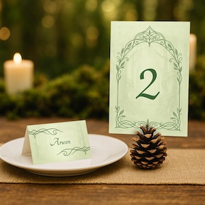 May include: A table setting featuring a light green table number card with the number 2 and a matching place card with the name "Arwen". A pine cone sits on a burlap table runner. A white plate is in the foreground.