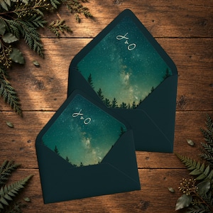 May include: Two dark teal envelopes with pointed flaps, each containing a card featuring a night sky with a starry galaxy, silhouettes of pine trees at the bottom, and the white cursive monogram J & O printed at the top center of the card.