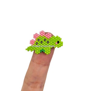 May include: A small, beaded stegosaurus in shades of green, pink, and black. The dinosaur has a row of pink plates along its back and is resting on a finger. The image includes the text "PDF Bead Pattern Stegosaurus".