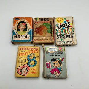 May include: Five vintage card game boxes are displayed, including "Old Maid," "Tree Spotter Cards," "Spots and Stripes," "Hearts and Crazy Eights," and a Pinocchio card game. The boxes feature colorful illustrations and text, showcasing classic game designs.