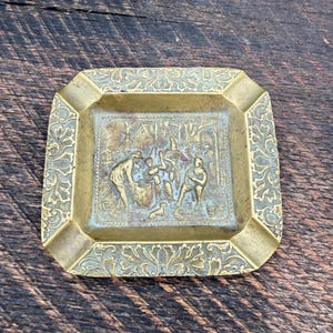 May include: A square brass ashtray featuring an embossed scene of figures in a rural setting at the center. The wide, sloped border is decorated with an intricate, repeating foliate scroll pattern. The metal has a warm, aged golden patina.