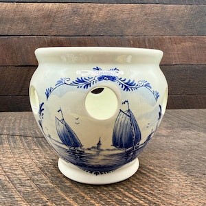 May include: A cream-colored ceramic bulbous vase featuring a hand-painted blue nautical scene with sailboats on water, a small building, and birds. The vessel has a wide rim and several circular cutouts around the upper body of the piece.