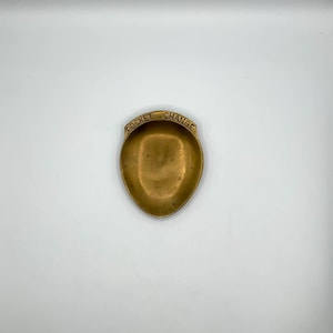 May include: A small, antique brass pocket change dish. The dish is oval-shaped with a slightly raised rim and a shallow interior. The words "POCKET CHANGE" are embossed on the top edge. The dish has a warm, golden hue.