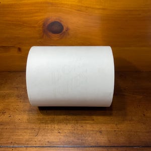 May include: A white roll of paper with the faint text "DOW JONES" printed on the surface. The roll is cylindrical and lies horizontally on a wooden surface. The paper appears to be a type of newsprint or similar material.