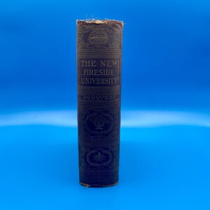 May include: A vintage book spine with the title "THE NEW FIRESIDE UNIVERSITY" in embossed gold lettering. The dark blue cover features ornate gold decorative patterns. The book is set against a bright blue background.