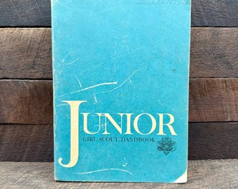 Könnte beinhalten: Ein blaugrünes Junior Girl Scout Handbuch mit einem großen cremefarbenen Buchstaben J und dem Text Junior Girl Scout Handbook auf dem Einband. Der Einband zeigt ein kleines Girl Scout Kleeblatt-Logo und handschriftlichen Text oben.
