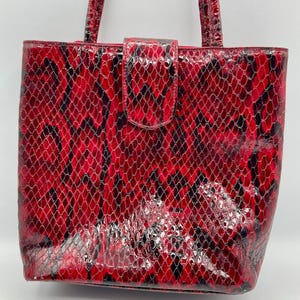 May include: A red and black snakeskin-patterned tote bag with a rectangular shape. The bag has two handles and a flap closure with a magnetic snap. The bag's exterior has a glossy finish.