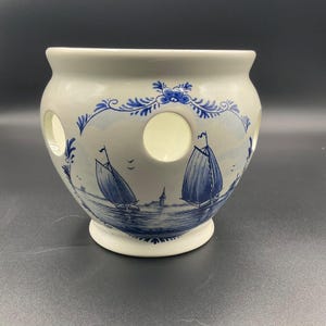 May include: White ceramic planter with blue sailboat and floral designs. The planter has a round shape with several circular openings and a pedestal base. The nautical scene features sailboats on the water. The rim is scalloped and decorated with blue floral accents.