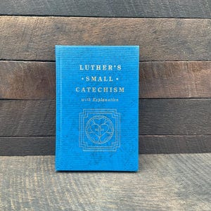 May include: A bright blue hardcover book titled Luther's Small Catechism with Explanation. The front cover features gold embossed text and a central Luther rose emblem enclosed within a square geometric border, set against a textured fabric cover.
