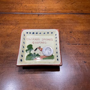 May include: A small, square ceramic dish with a painted scene of a house and trees, and the words "Colorado Springs Colorado." The dish has a cream-colored base, a red border, and blue dot accents. A quarter coin rests inside the dish.