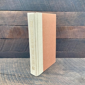 May include: A hardcover book titled "THE LANGUAGE OF THE HEART" with a beige spine and a peach-colored cover. The title is printed vertically in gold lettering on the spine. The book is standing upright against a dark wood background.