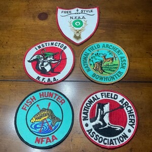 May include: A collection of five embroidered patches with various designs and text. The patches feature images of a deer, squirrel, fish, and archery-related graphics. The patches are in various colors, including red, green, and blue.