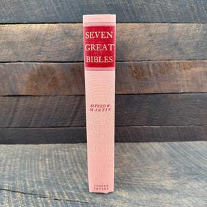 May include: A light pink hardcover book with a red rectangular panel on the spine. The text in white reads SEVEN GREAT BIBLES and ALFRED W. MARTIN, with COOPER SQUARE at the bottom. The book has a textured fabric cover and a vertical orientation.