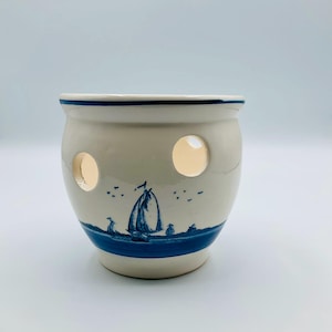 May include: A ceramic candle holder with a nautical theme. The off-white holder features a blue sailboat design and three circular cutouts. A blue band and trim complete the design. The candle holder is approximately 4 inches tall.
