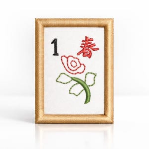 May include: A framed Mahjong tile with the number 1 in black, a red Chinese character, and a red and green floral design. The frame is a light brown colour.