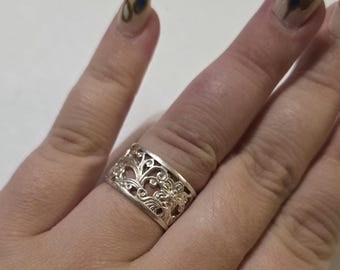 May include: A silver ring with an openwork floral design. The ring features intricate details of flowers and swirling vines. The ring is worn on a finger, showcasing its delicate craftsmanship and elegant style. The ring is a piece of jewelry.