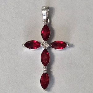 May include: A silver-colored pendant in the shape of a cross, featuring five marquise-cut, deep red gemstones. The gemstones are set in a silver metal frame, with a small loop at the top for a chain.