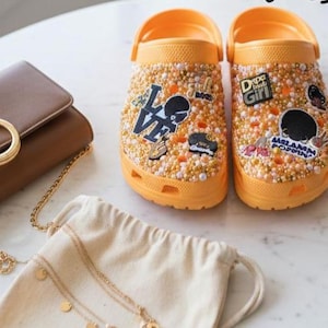 Op de afbeelding: Een paar oranje klompen versierd met kleine, glinsterende kralen en decoratieve patches. De patches tonen tekst en afbeeldingen. Een bruine handtas met een gouden ketting en een kleine gouden ketting zijn ook zichtbaar.