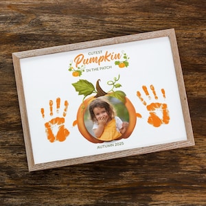 May include: A framed print with a wooden frame featuring a child's photo inside an orange pumpkin illustration. Orange handprints flank the pumpkin, with the text "Cutest Pumpkin in the Patch" and "Autumn 2025".
