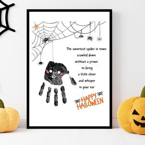 Pode incluir: Uma impressão emoldurada com tema de Halloween com um design de aranha de impressão manual e o texto "The sweetest spider in town...Happy Halloween." Abóboras decorativas e uma teia de aranha também são visíveis.