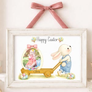 May include: A framed Easter-themed print featuring a baby in an egg-shaped frame being pulled in a wooden wagon by a cartoon bunny. The print includes the text "Happy Easter" and the name "Olivia". The frame is white and is suspended by a pink ribbon.