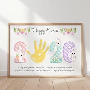 May include: A framed print with the words "Happy Easter" above the numbers "2020". The "2"s are decorated with bunny illustrations. A yellow handprint is in the center. The text below reads "A tiny hand pressed down with care..."