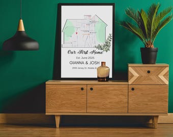 May include: A framed print with a green and white map of a neighborhood, with the text "Our First Home" and "Est. June 2025" above the map. Below the map, the text reads "GIANNA & JOSH" and "2930 Jersey Dr. Mobile Al". The print is sitting on a wooden shelf with a small plant in a black pot to the right.