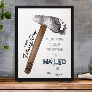 May include: A framed print featuring a hammer design and a footprint, with the text "Happy Father's Day 2026." The message reads "When it comes to being the best dad, you nailed it!" with the name "Ethan." The frame is black.