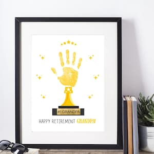 Peut inclure: Impression encadrée avec un motif de trophée d'empreinte de main dorée. L'impression présente le texte "#1 GRANDPA" et "HAPPY RETIREMENT GRANDPA!" en noir. Des étoiles dorées ornent l'empreinte, sur fond blanc.